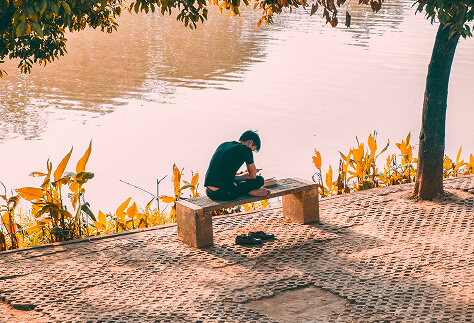 A photograph of a man sitting and reading on a bench at the riverside under a tree on a sunny day.